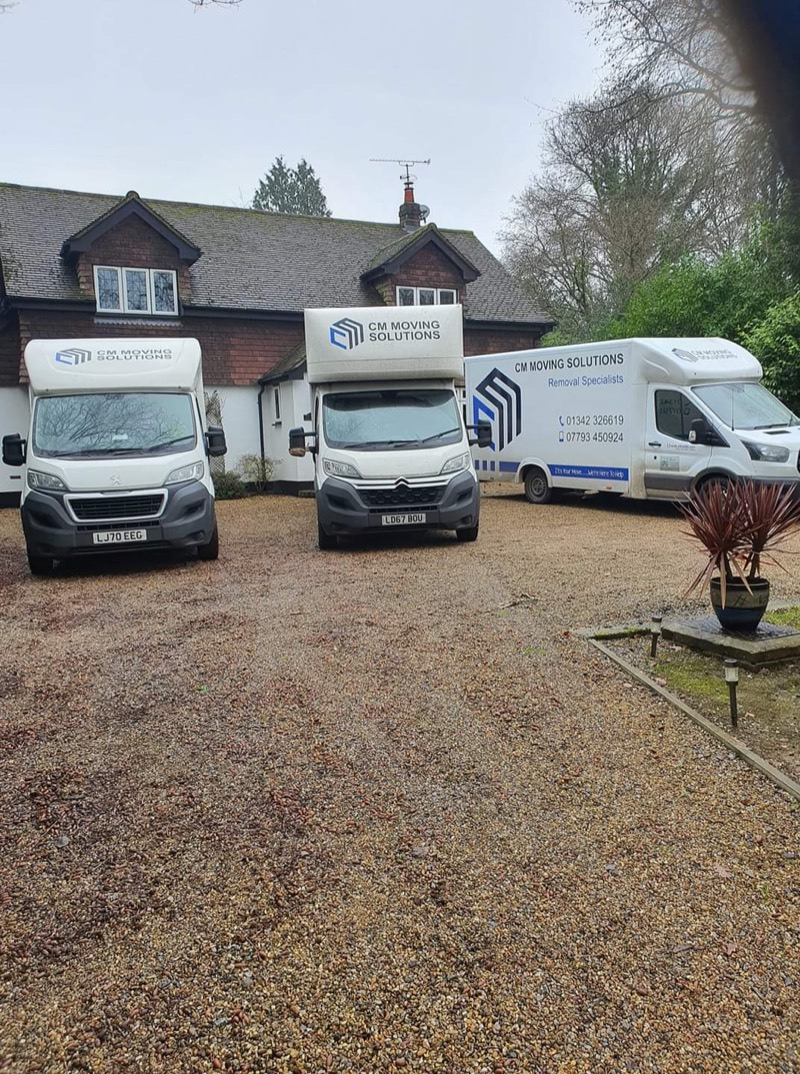 Three CM Moving Solutions removal vans parked on a gravel driveway in front of a house